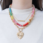 Model wearing stacked necklaces. One is a dainty gold necklace with a bar pendant, one is a medium sized multi-colored beaded necklace and one is a large accent gold chained charm necklace with a large gold heart pendant with a diamond at its center.