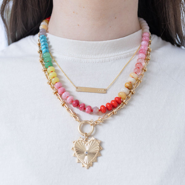 Model wearing stacked necklaces. One is a dainty gold necklace with a bar pendant, one is a medium sized multi-colored beaded necklace and one is a large accent gold chained charm necklace with a large gold heart pendant with a diamond at its center.