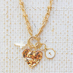 Picture of gold toggle chain with a textured gold heart pendant, a white cross charm and a white disc charm with a gold cross in the center.