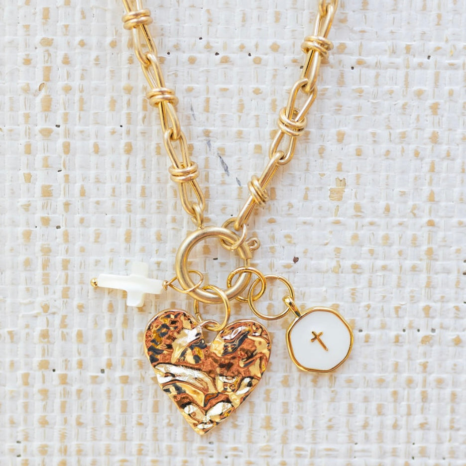 Picture of gold toggle chain with a textured gold heart pendant, a white cross charm and a white disc charm with a gold cross in the center.