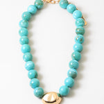 Chunky turquoise beaded necklace with a large oval gold bead and gold clasp on a white background