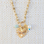 Picture of gold toggle chain with large gold heart pendant with a diamond at the center. Necklace also has a light teal bead, a fresh water bead and an evil eye attached.