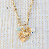 Picture of gold toggle chain with large gold heart pendant with a diamond at the center. Necklace also has a light teal bead, a fresh water bead and an evil eye attached.