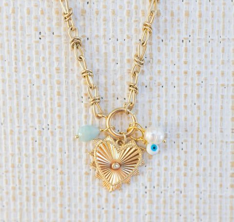 Picture of gold toggle chain with large gold heart pendant with a diamond at the center. Necklace also has a light teal bead, a fresh water bead and an evil eye attached.