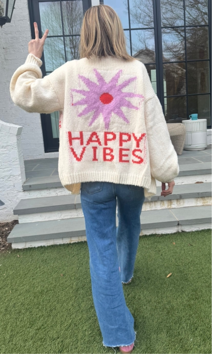 Picture of a woman walking away, holding up her hand in a peace sign, wearing an oversized cream colored sweater with a large purple sunburst with a red center and the words Happy Vibes in red at the bottom.