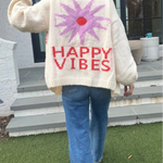 Picture of a woman walking away, holding up her hand in a peace sign, wearing an oversized cream colored sweater with a large purple sunburst with a red center and the words Happy Vibes in red at the bottom.
