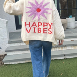 Picture of a woman walking away, holding up her hand in a peace sign, wearing an oversized cream colored sweater with a large purple sunburst with a red center and the words Happy Vibes in red at the bottom.