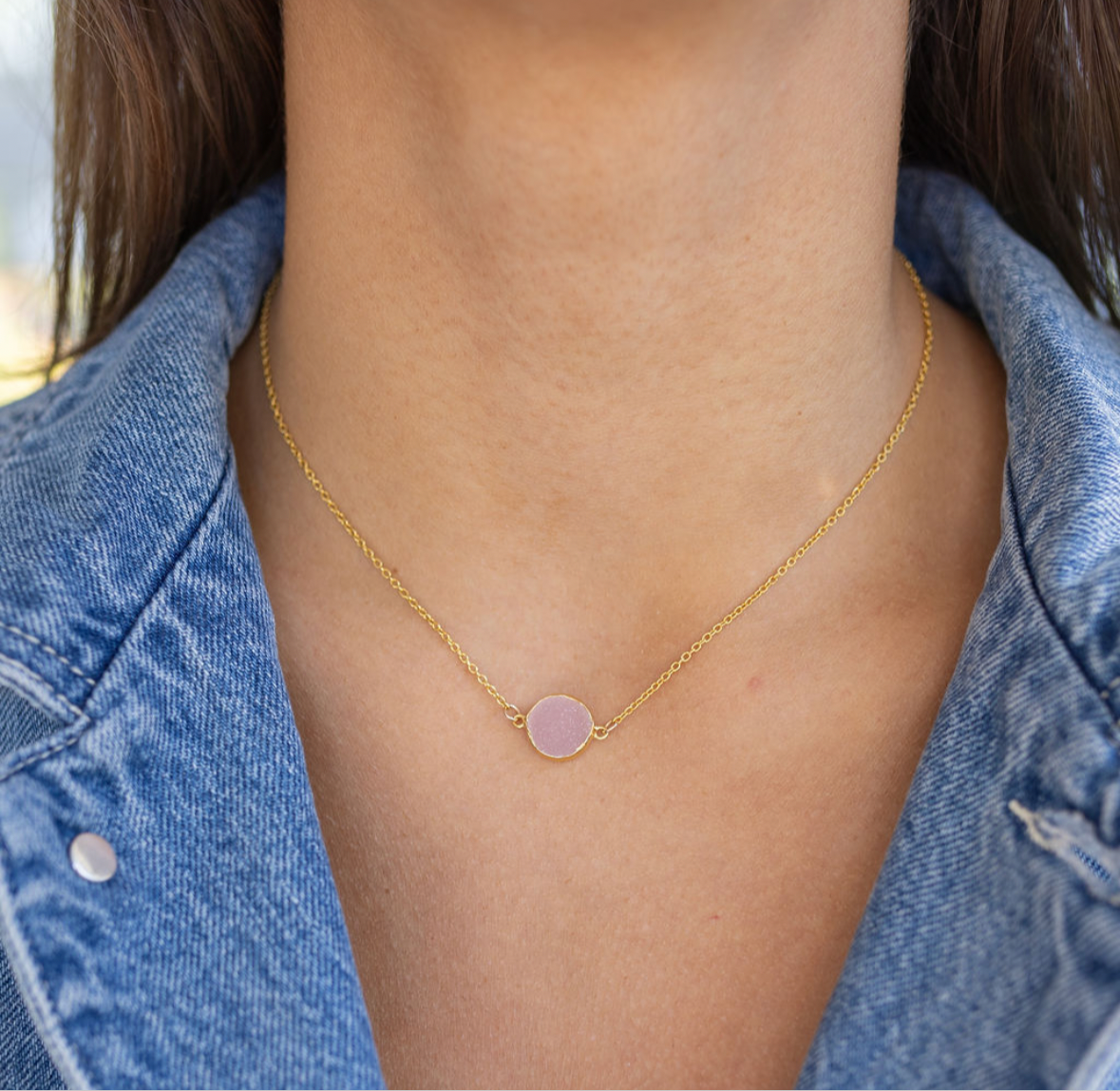 Gold necklace with a pink pendant worn by a person in a denim jacket.