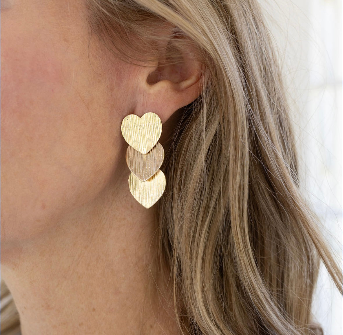 A woman with blonde hair wearing a pair of triple heart brushed gold dangling earrings.