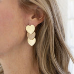 A woman with blonde hair wearing a pair of triple heart brushed gold dangling earrings.