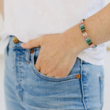 Picture of a woman with her hand in her pocket. She is wearing a beaded bracelet in greens, corals, white and some browns.