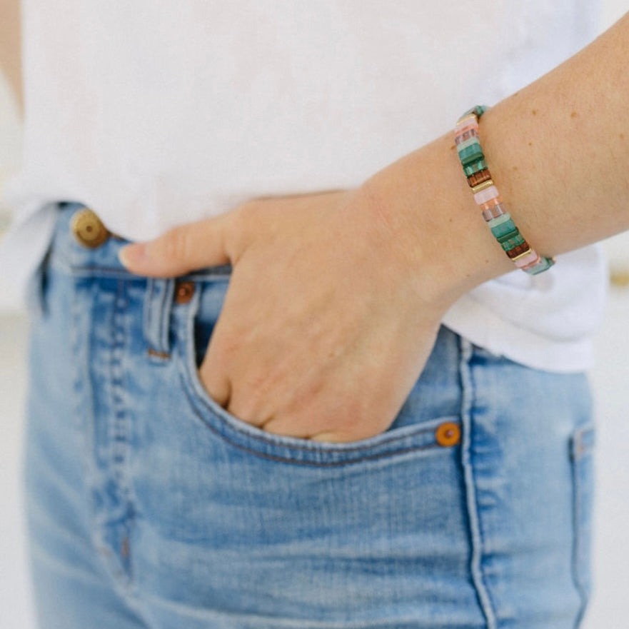 Picture of a woman with her hand in her pocket. She is wearing a beaded bracelet in greens, corals, white and some browns.