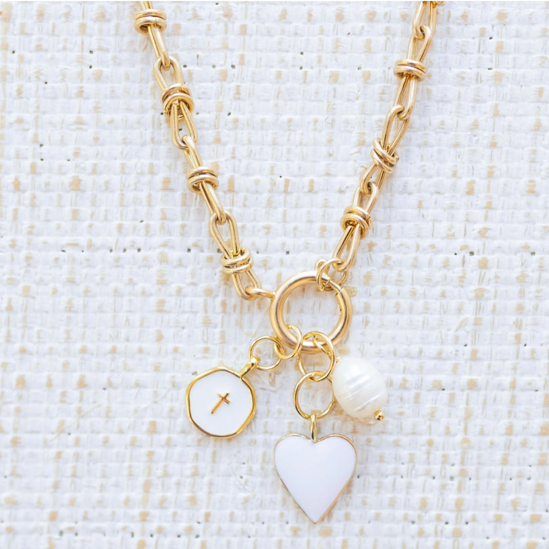 Picture of gold toggle chain with a white heart charm, white disc charm with a cross at the center and a fresh water pearl charm.