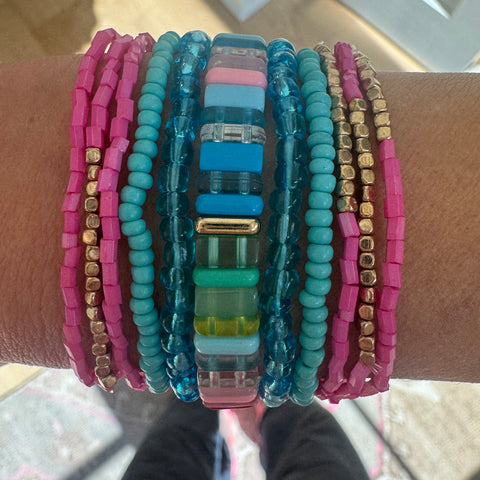 Picture of a woman's wrist displaying a beautiful set of bracelets. Six pink and gold small bead bracelets, 2 solider blue bracelets, 2 clear blue bracelets and one thicker beaded bracelet made of rectangular beads in blues, greens and pinks.
