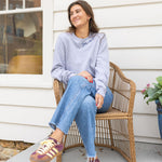 Woman sitting in a chair on a porch wearing a gray sweatshirt with blue stitching that says  "Joie De Vivre"