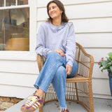 Woman sitting in a chair on a porch wearing a gray sweatshirt with blue stitching that says  "Joie De Vivre"