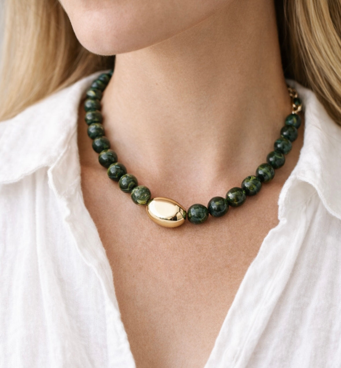 Green beaded necklace with a large central gold bead and gold clasp worn by a person in a white shirt.