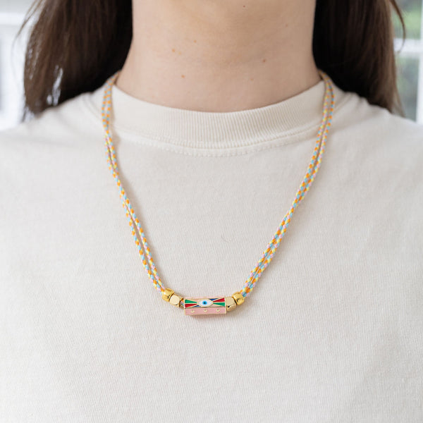 Model wearing a braided cord necklace with two strands. Necklace has gold beads and rectangle evil eye pendant at the center.