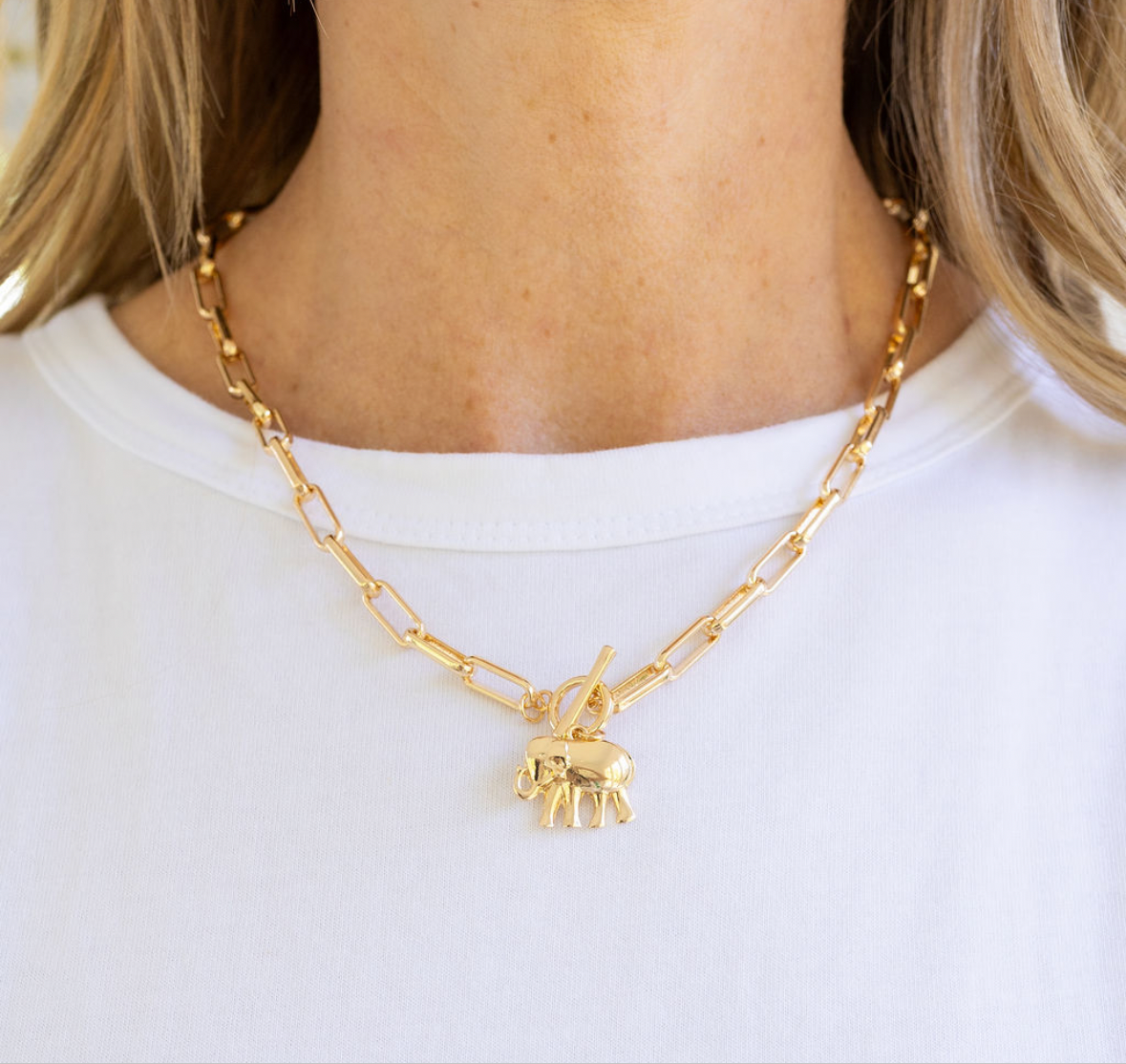 Woman wearing a large gold rectangle chain necklace with a toggle clasp and a gold elephant pendant.