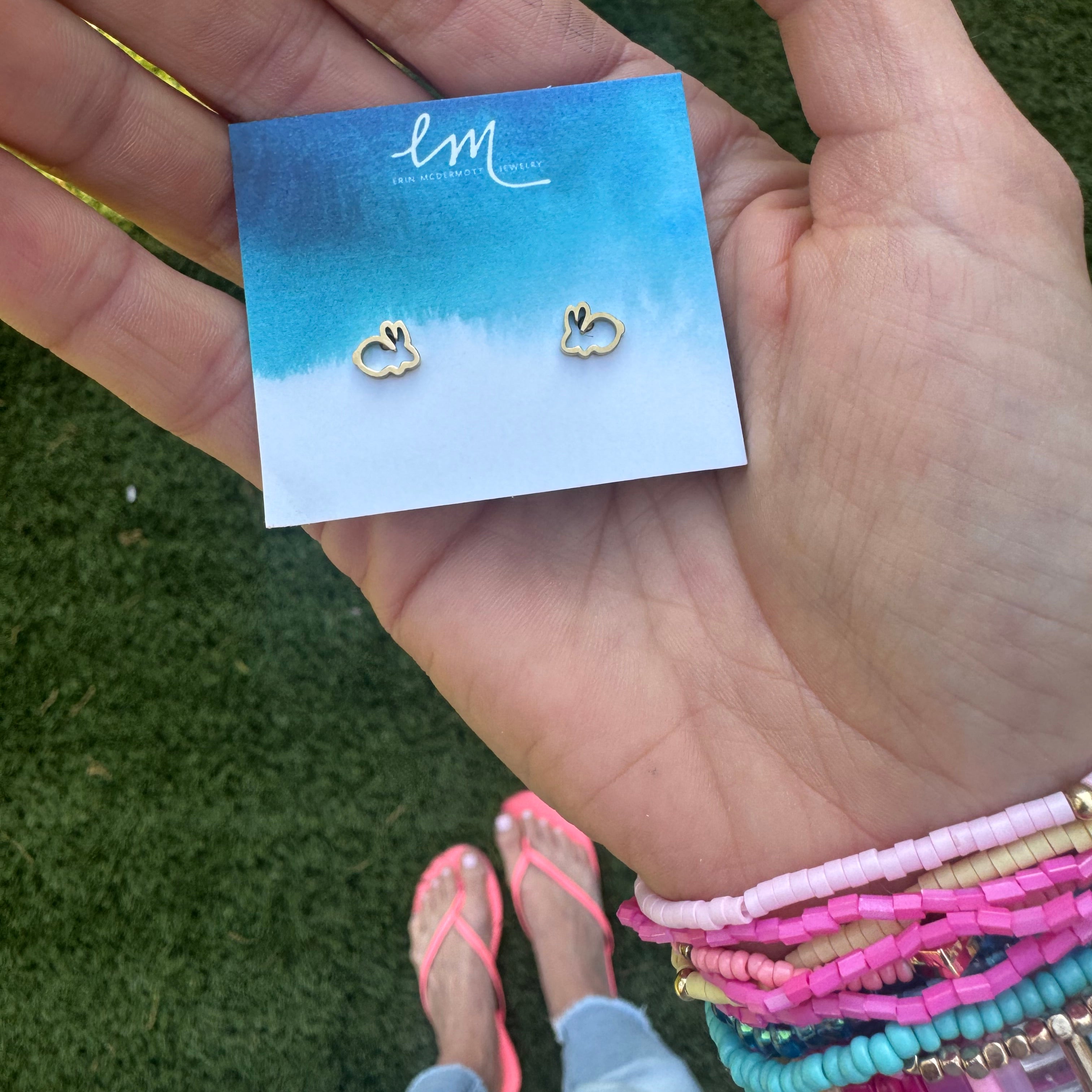Picture of a woman's hand with many colorful beaded bracelets on her wrist holding an earring holder displaying two delicate gold outline bunny earrings.