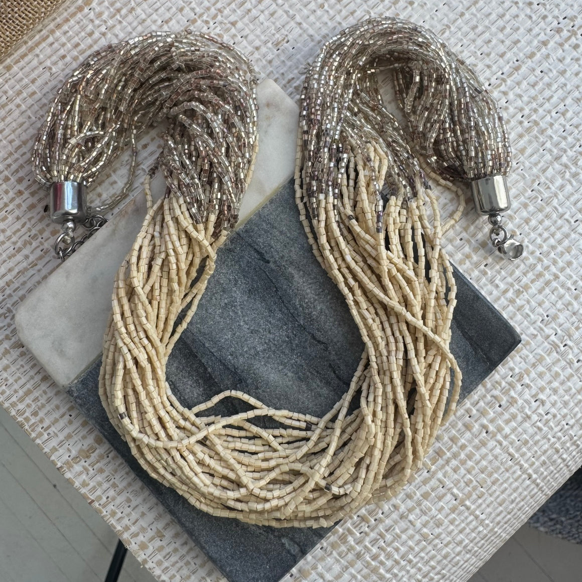 Picture of a statement necklace made of many strands of cream colored seed beads. Strands begin with clear tan beads, have a tan section in the middle and end with clear tan beads.