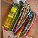 Woman's wrist displaying a cluster of 14 colorful bracelets stacked with a larger rectangle beaded stretch bracelet.