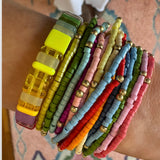 Woman's wrist displaying a cluster of 14 colorful bracelets stacked with a larger rectangle beaded stretch bracelet.