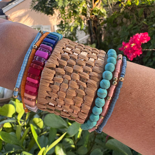 Picture of arm with 5 bracelets and one tan, woven cuff. Beaded bracelets are turquoise, coral, multicolored, blue and orange.