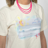 Screenshot of a woman wearing a t shirt displaying a beach scene and wearing bright pink beaded necklaces.