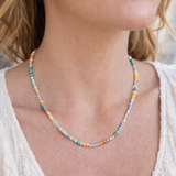 Close-up of a blue, yellow and clear colorful beaded necklace on a person wearing a light-colored top.