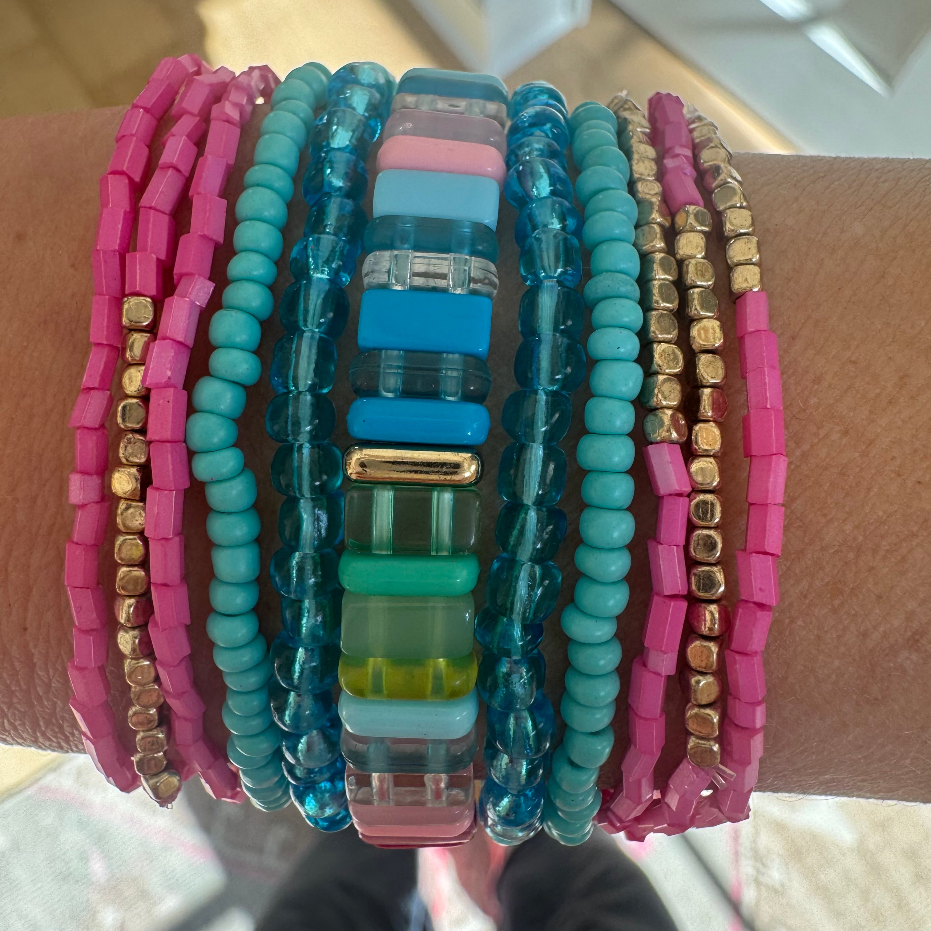 Picture of a woman's wrist displaying a beautiful set of bracelets. Six pink and gold small bead bracelets, 2 solider blue bracelets, 2 clear blue bracelets and one thicker beaded bracelet made of rectangular beads in blues, greens and pinks.