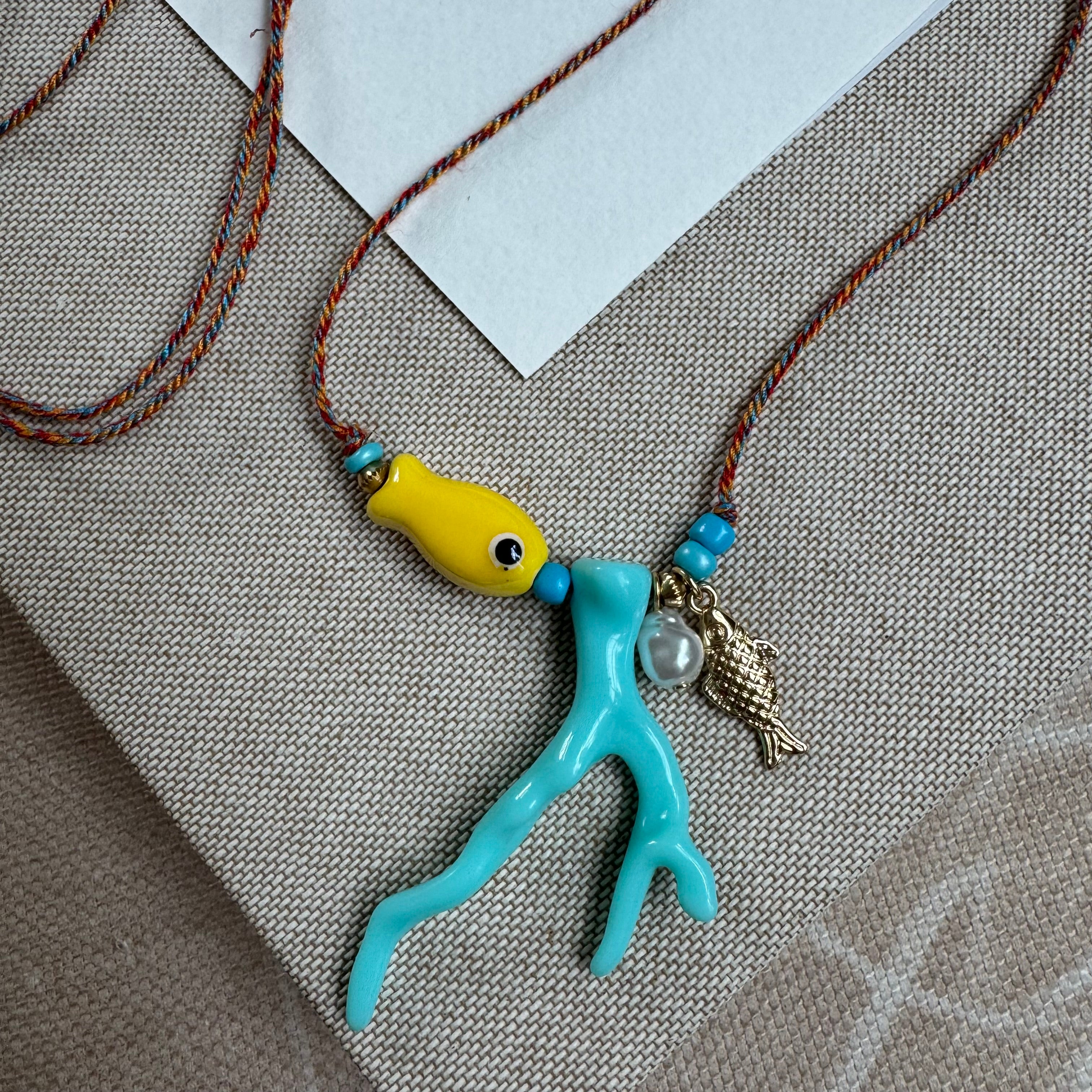 Picture of a woven string necklace with a yellow fish bead, a light blue coral bead, a fresh water pearl and a small gold fish pendant.
