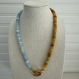 Picture of a bead necklace that is half light blue and half dark amber yellow. Necklace has a gold carabiner clasp.