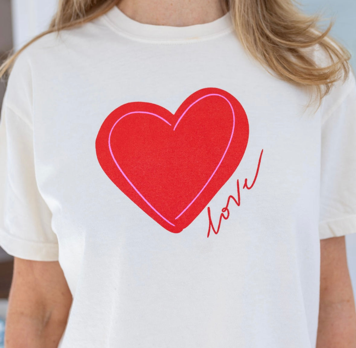 Woman wearing a a beige Tshirt with a red heart and script that reads "love".