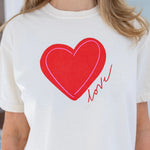 Woman wearing a a beige Tshirt with a red heart and script that reads "love".