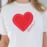 Woman wearing a a beige Tshirt with a red heart and script that reads "love".