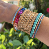 Picture of arm with 5 bracelets and one tan, woven cuff. Beaded bracelets are turquoise, coral, multicolored, blue and orange.