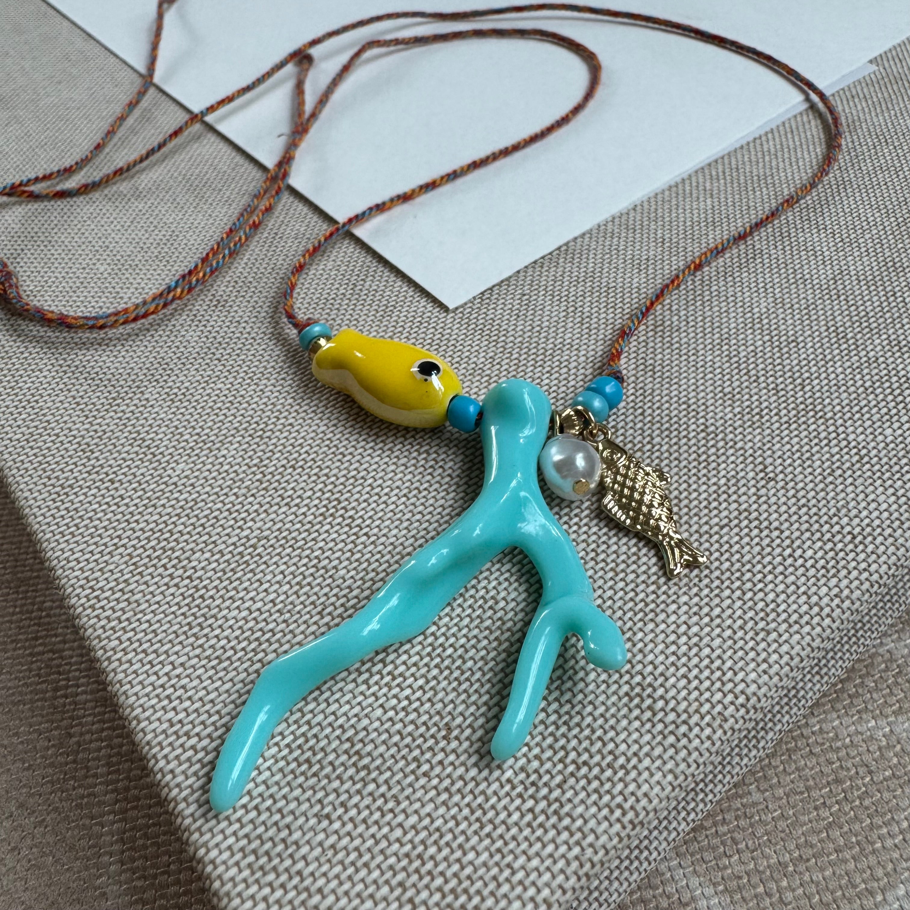 Picture of a woven string necklace with a yellow fish bead, a light blue coral bead, a fresh water pearl and a small gold fish pendant.