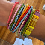 Woman's wrist displaying a cluster of 14 colorful bracelets stacked with a larger rectangle beaded stretch bracelet.