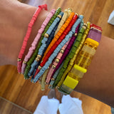 Woman's wrist displaying a cluster of 14 colorful bracelets stacked with a larger rectangle beaded stretch bracelet.