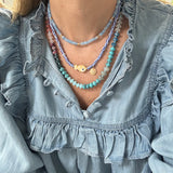 Model in a denim ruffle shirt displaying two necklaces, one a dainty blue bead necklace with a small white disc pendant. Another necklace is made of blue and white striped beads with a tan polka dot fish bead at its center and a small gold disc pendant. The third is a bead necklace with turquoise beads, ombre rose colored beads and various cream opaque beads.