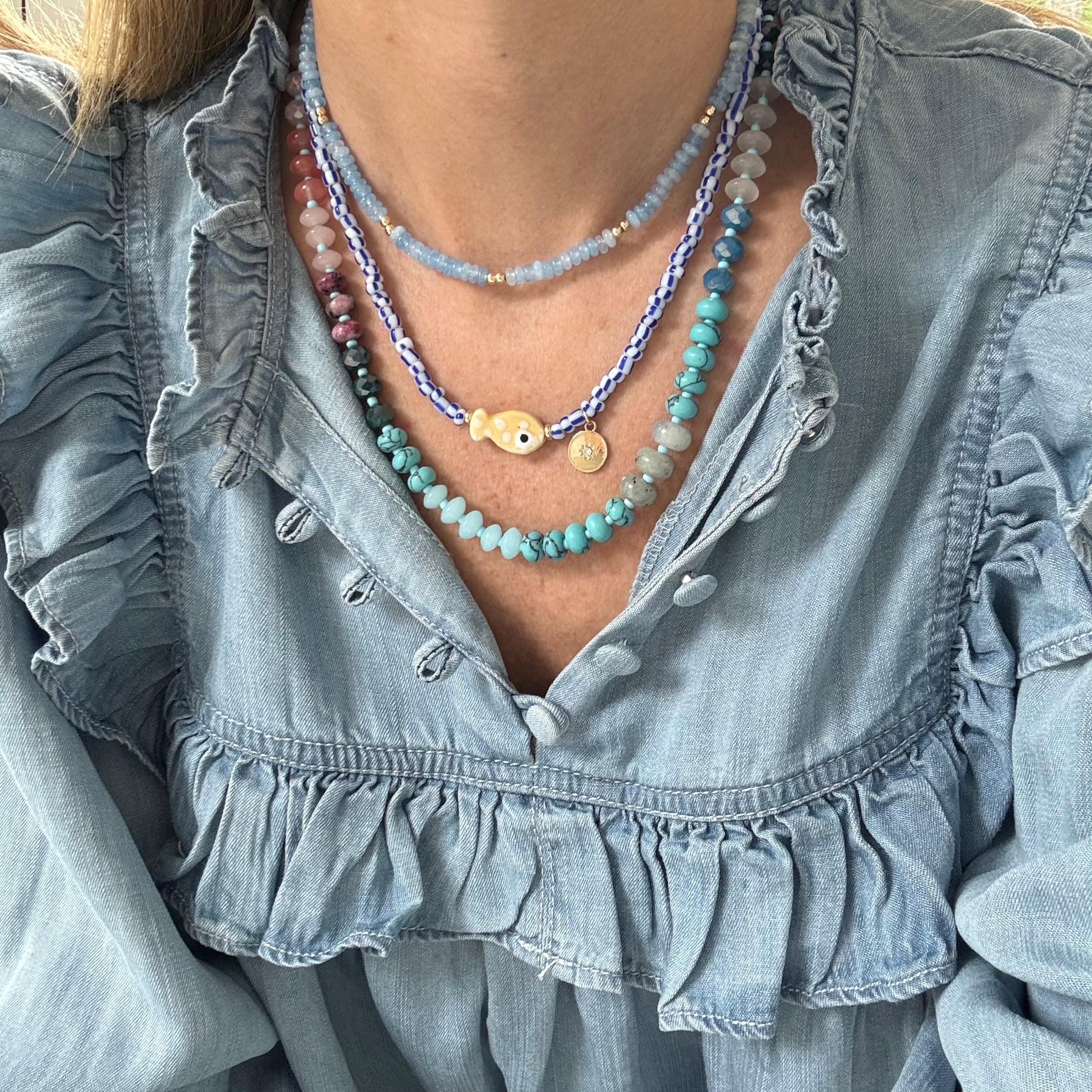 Model in a denim ruffle shirt displaying two necklaces, one a dainty blue bead necklace with a small white disc pendant. Another necklace is made of blue and white striped beads with a tan polka dot fish bead at its center and a small gold disc pendant. The third is a bead necklace with turquoise beads, ombre rose colored beads and various cream opaque beads.