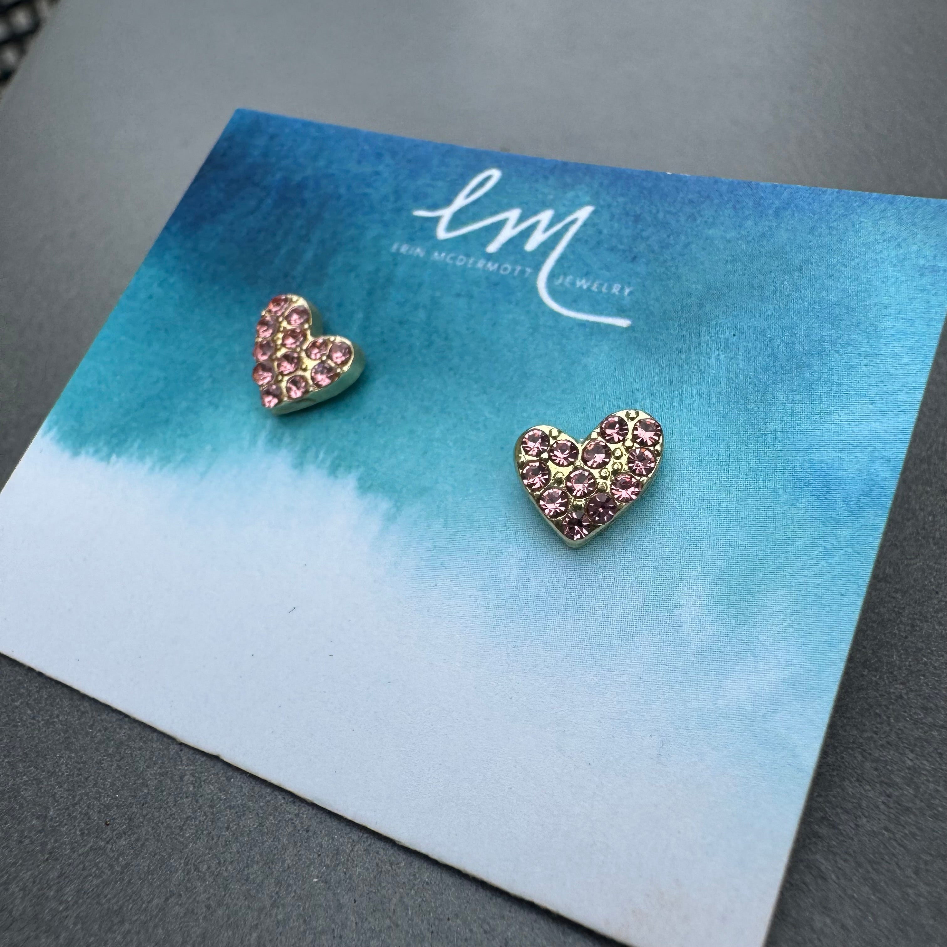 Heart-shaped earrings with pink stones on a blue card with 'EM' branding.