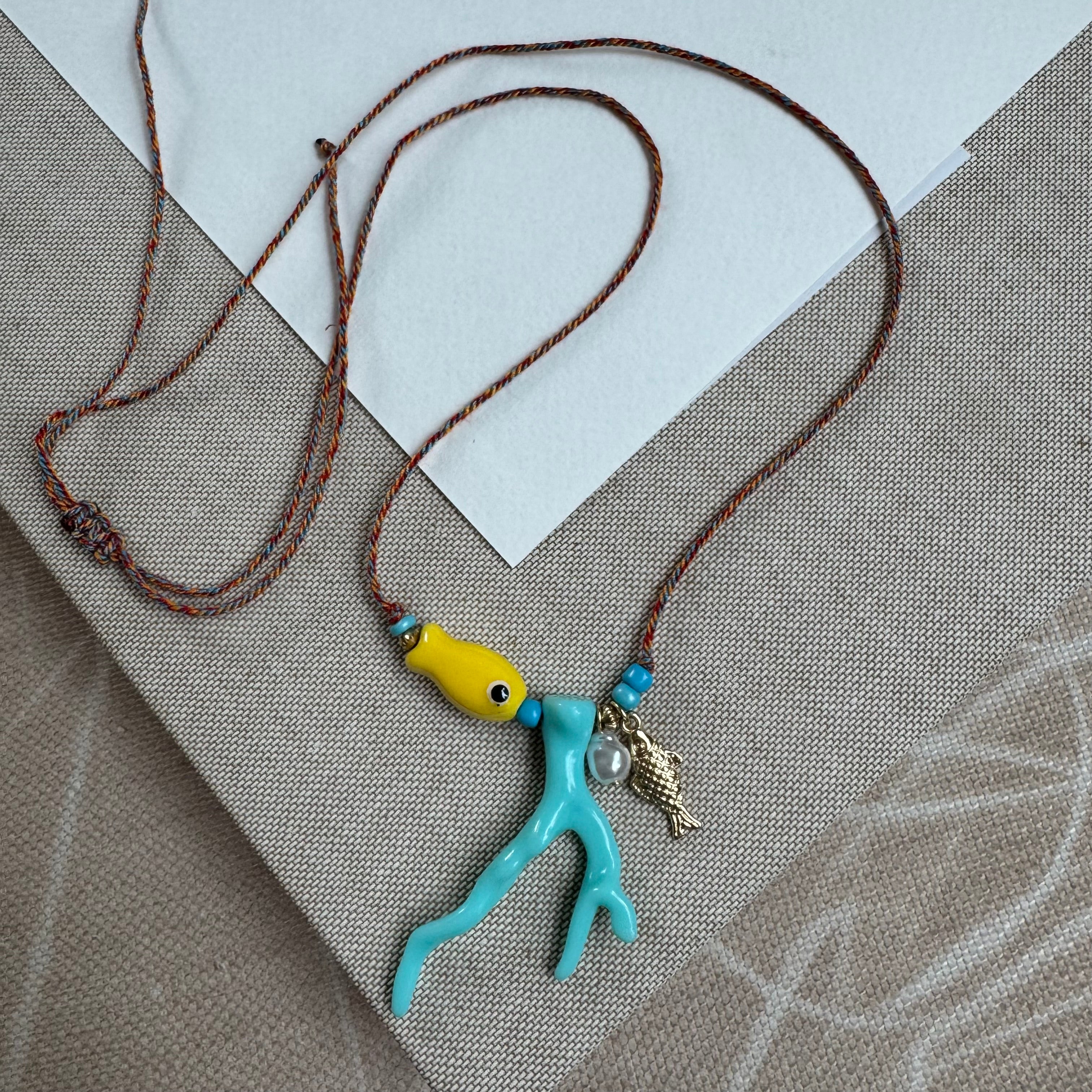 Picture of a woven string necklace with a yellow fish bead, a light blue coral bead, a fresh water pearl and a small gold fish pendant.