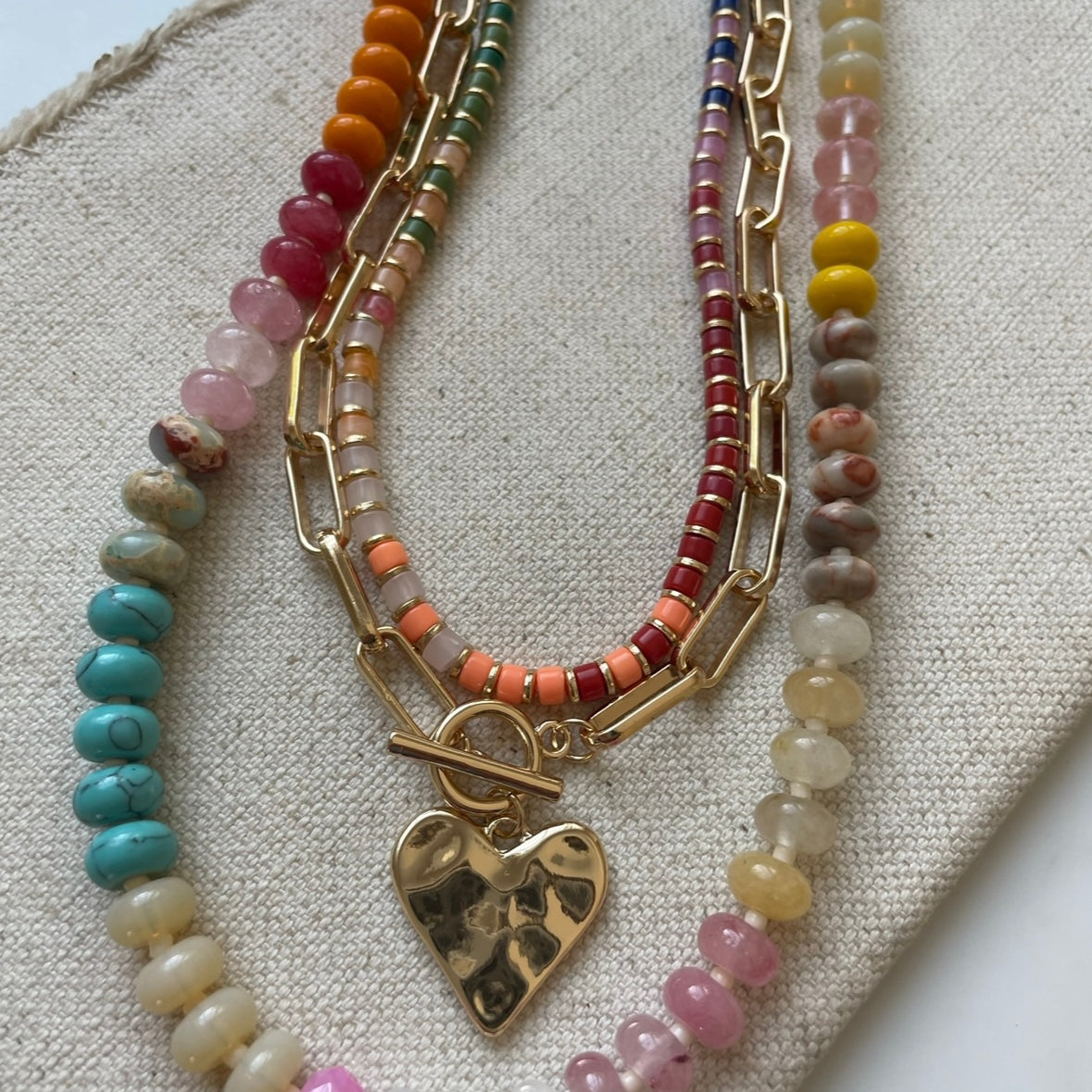 Set of three necklaces made up of small colorful bead necklace, a gold chain link with hammered gold heart pendant and a larger colorful beaded necklace.