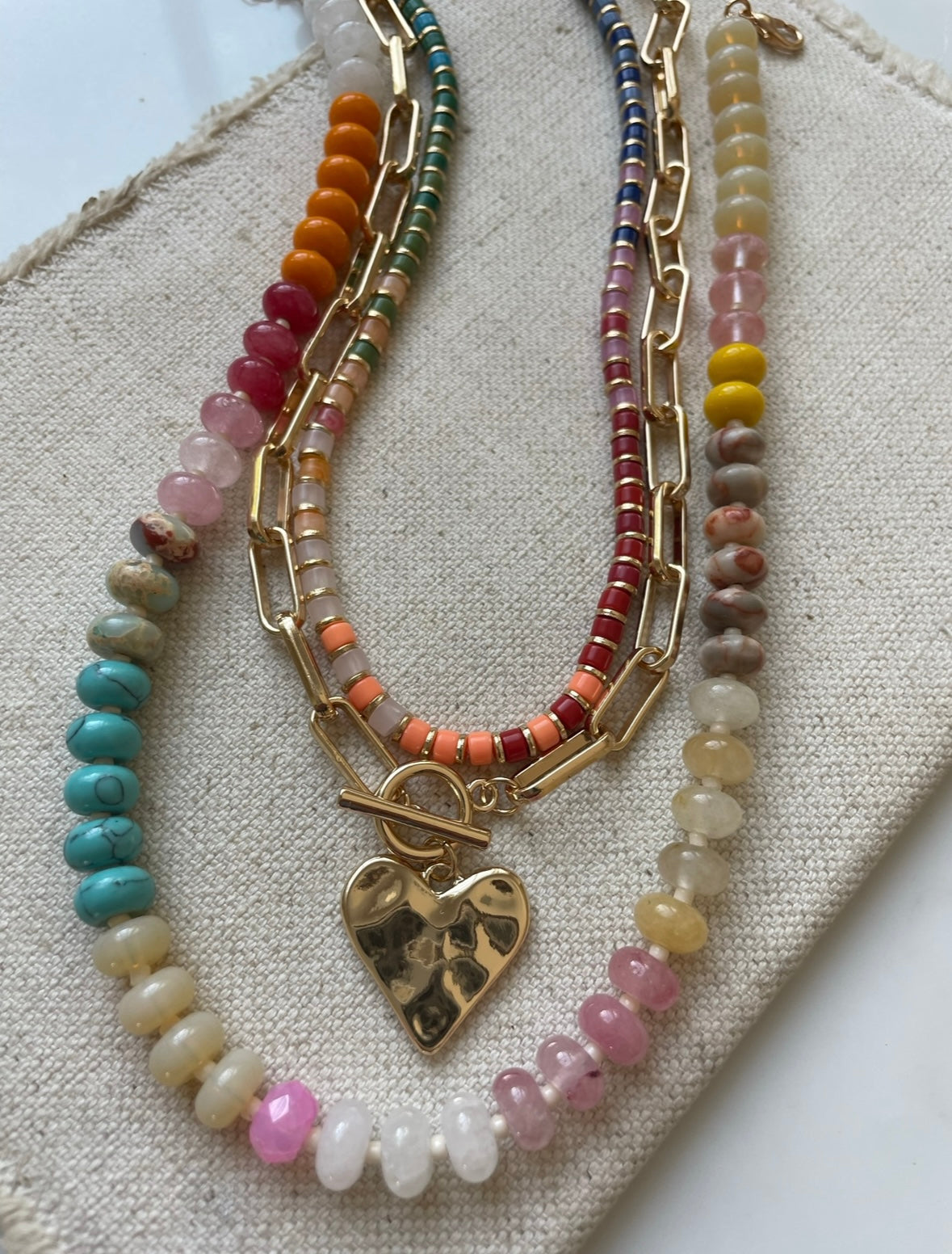 Set of three necklaces made up of small colorful bead necklace, a gold chain link with hammered gold heart pendant and a larger colorful beaded necklace.