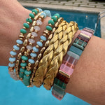 Picture of wrist with 3 beaded bracelets in shades of blue, two small gold bead bracelets, two woven larger gold bracelets and one multicolored, purple and pink beaded bracelet.