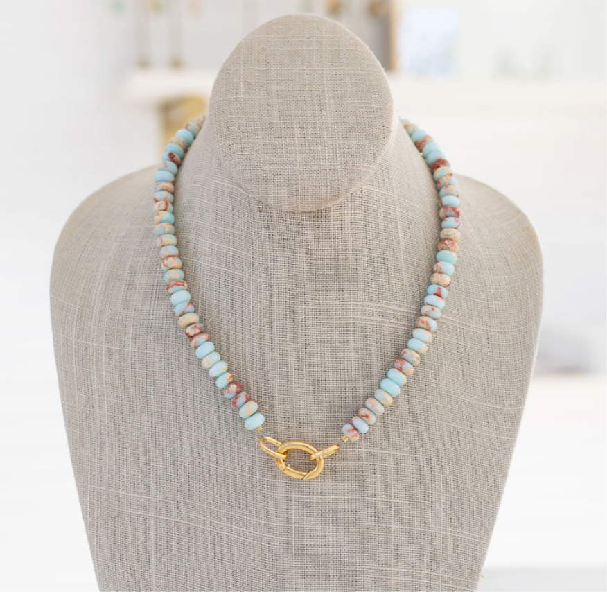 A semi-precious stone necklace with shades of baby blue, rust, and sand, featuring a gold carabiner clasp, displayed on a mannequin.