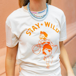 Woman wearing a cream shirt with an orange print of a cowgirl with a lasso and the text "Stay Wild" with multiple blue beaded necklaces