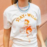 Woman wearing a cream shirt with an orange print of a cowgirl with a lasso and the text "Stay Wild" with multiple blue beaded necklaces